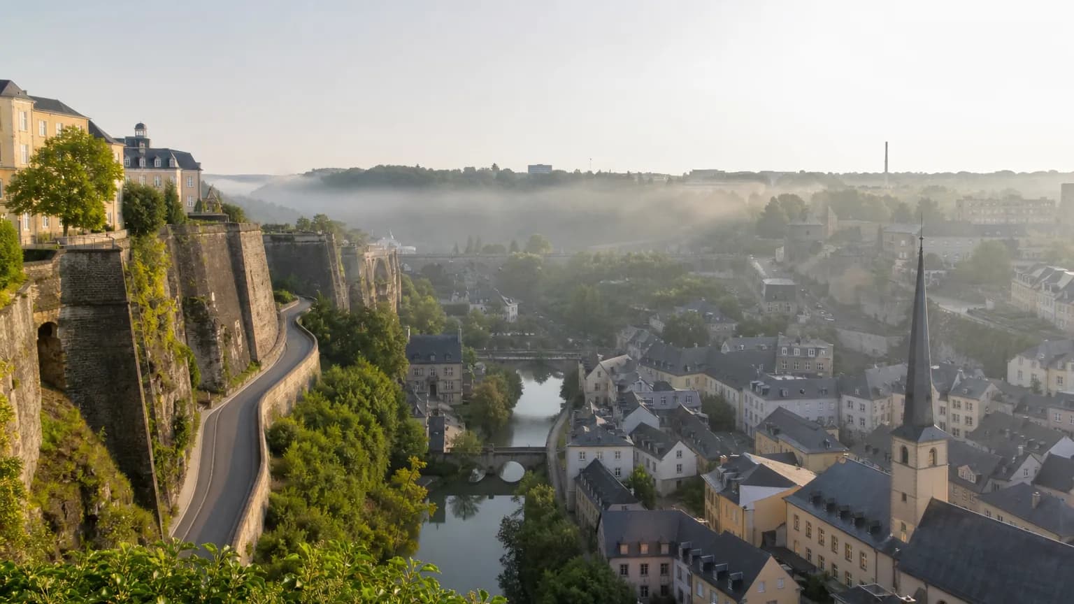Une vieille ville européenne en fond de vallée, ses toits d'ardoise, ses ponts de pierre et ses remparts, dans la brume du matin.