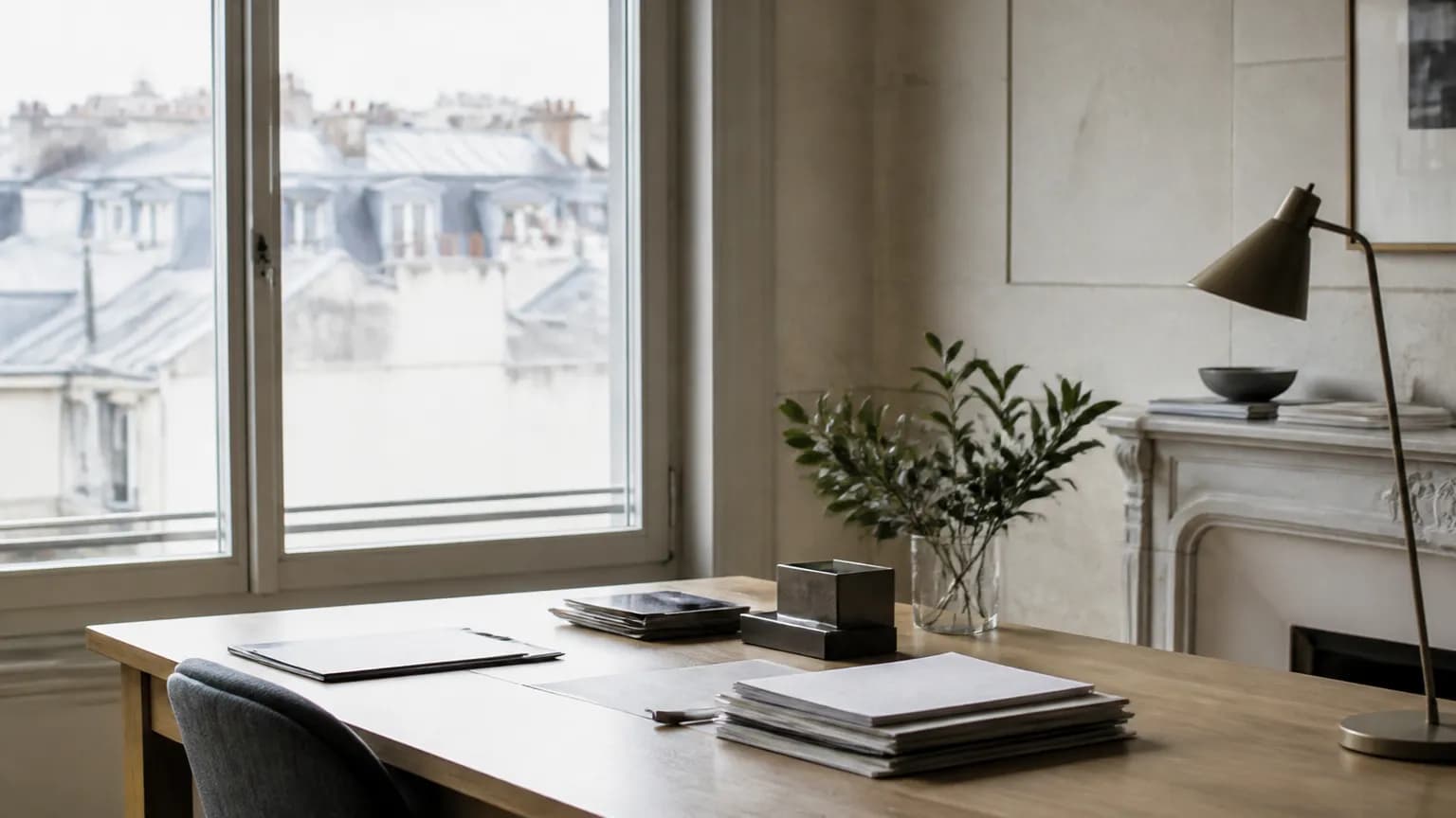 Un bureau clair face à une fenêtre ouverte sur des toits parisiens, documents et plante posés dessus.